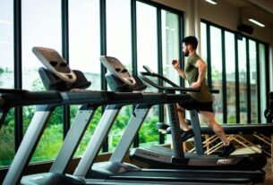 Young man workouts on treadmill in modern gym with large windows and natural light.