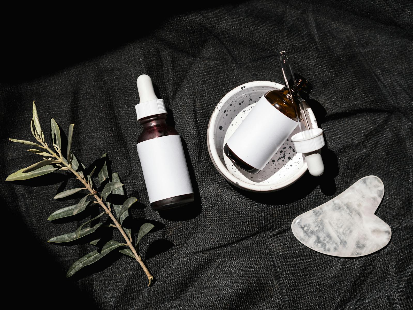 Flat lay of skincare serums and gua sha on black fabric with leaves.